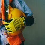 Close-up of a construction worker holding a hardhat and wearing PPE, including gloves and goggles.